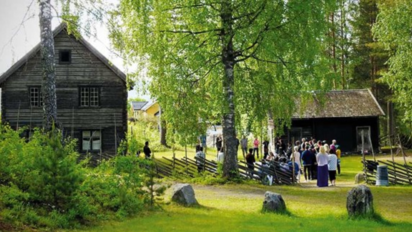 Turister på gårdstun mellom gamle hus og skigard. 