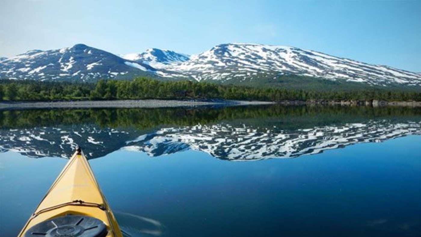 Kajakk på stille vann og snødekte fjell speiles i vannet. 