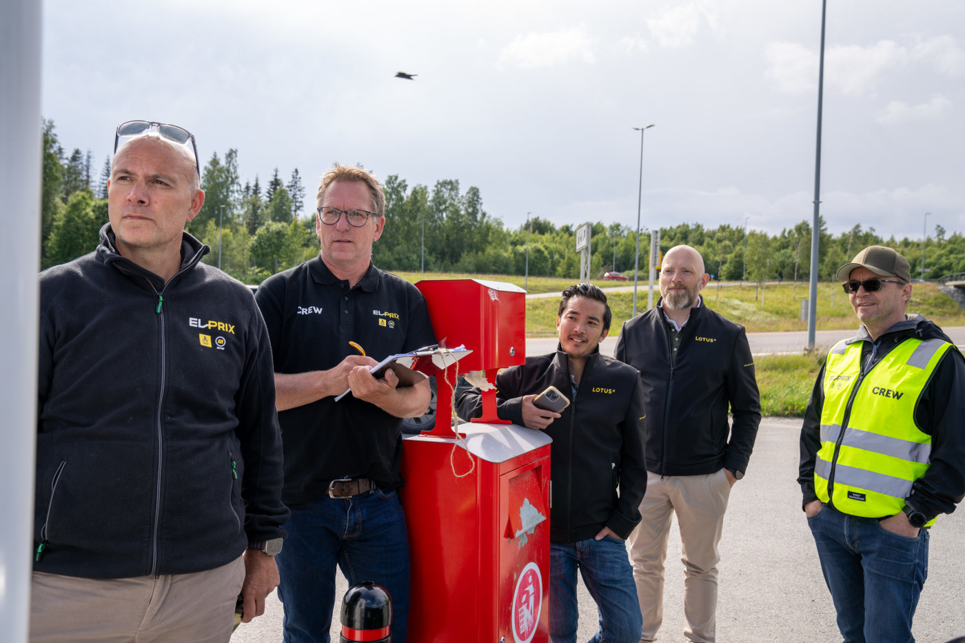 En gruppe menn står rundt en ladestolpe og følger nøye med.