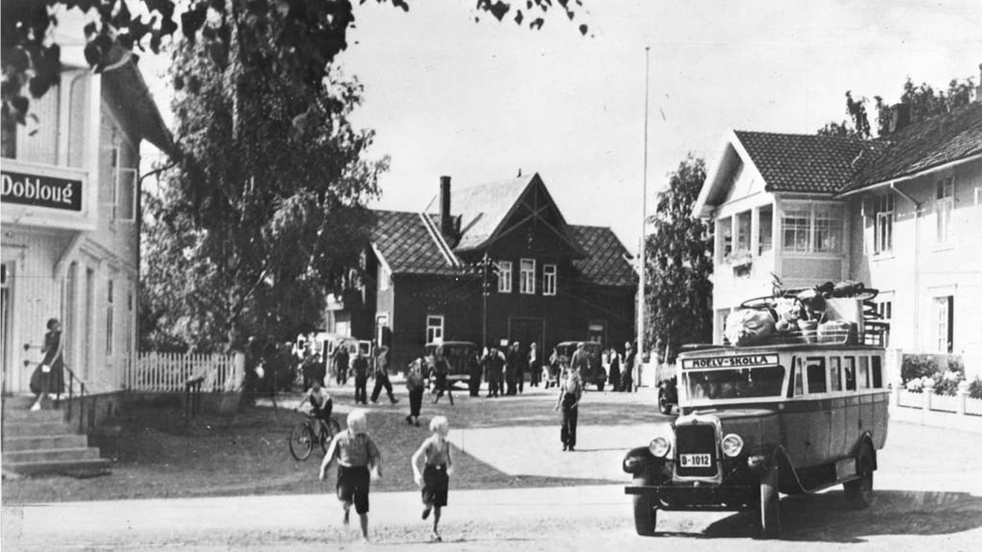 Historisk bilde av liten buss som kjører over et torg i Norge. Foran bilen løper to små barn.
