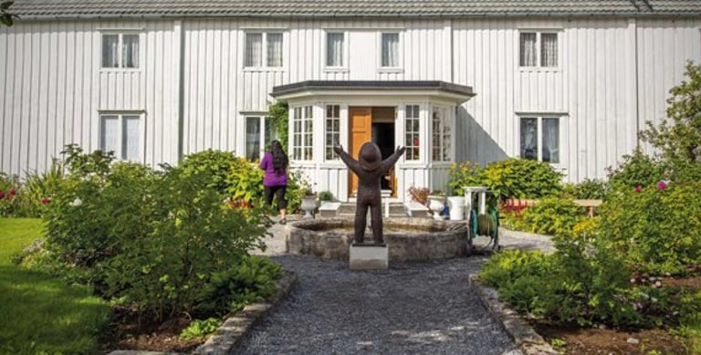 Gammel gård med grusbelagt inngangsparti og skulptur i front. 