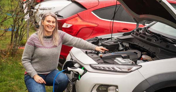 Dame sitter foran bilen med åpent panser. Smiler og peker på batteriet