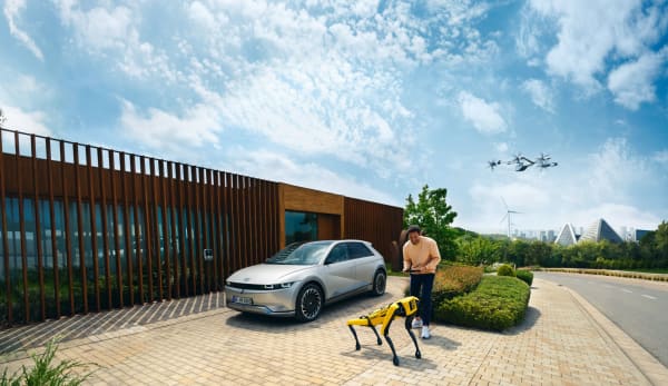Mann leker med en robot utenfor et hus. Bak ham står en elbil og lader mens en flyvende bil flyr over hodet hans.