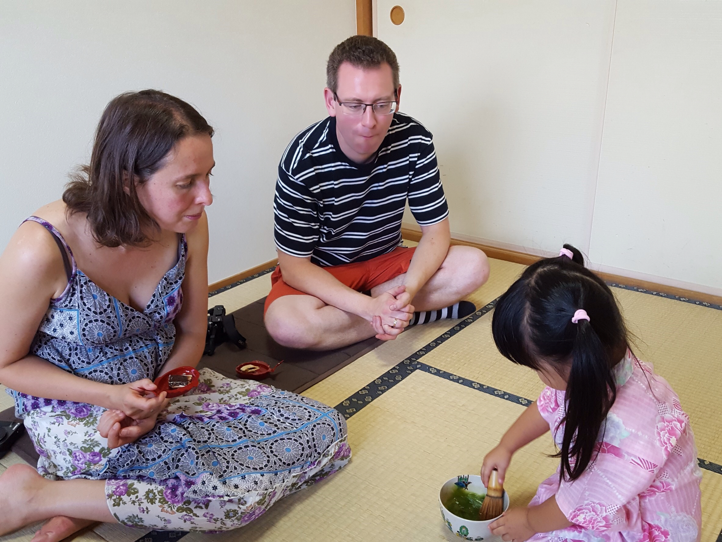 A couple sits on tatami mats while a young girl plays with a small bowl.