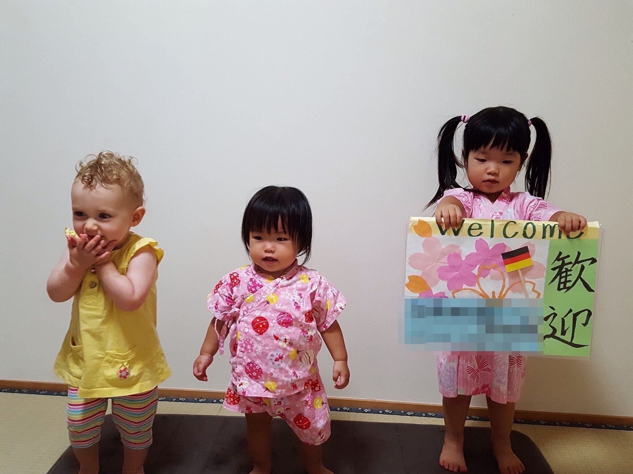 Three toddlers stand against a white wall, and one girl holds a Welcome sign.
