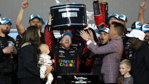 A man holds the Harley J. Earl Daytona 500 trophy over his head with a woman, a child, and several other men around him.