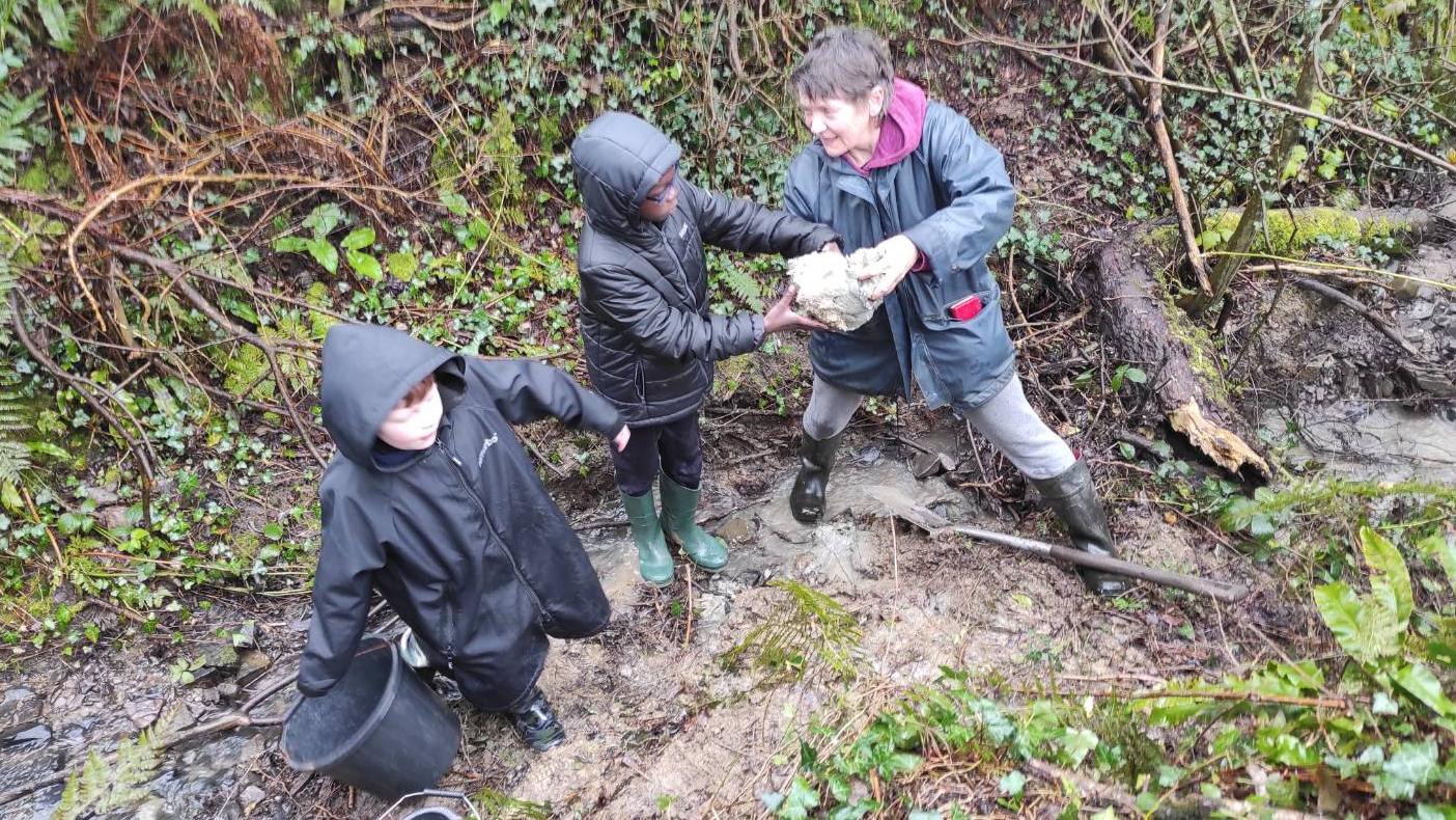Damp looking adult digging for clay by a stream, helped by two young students!