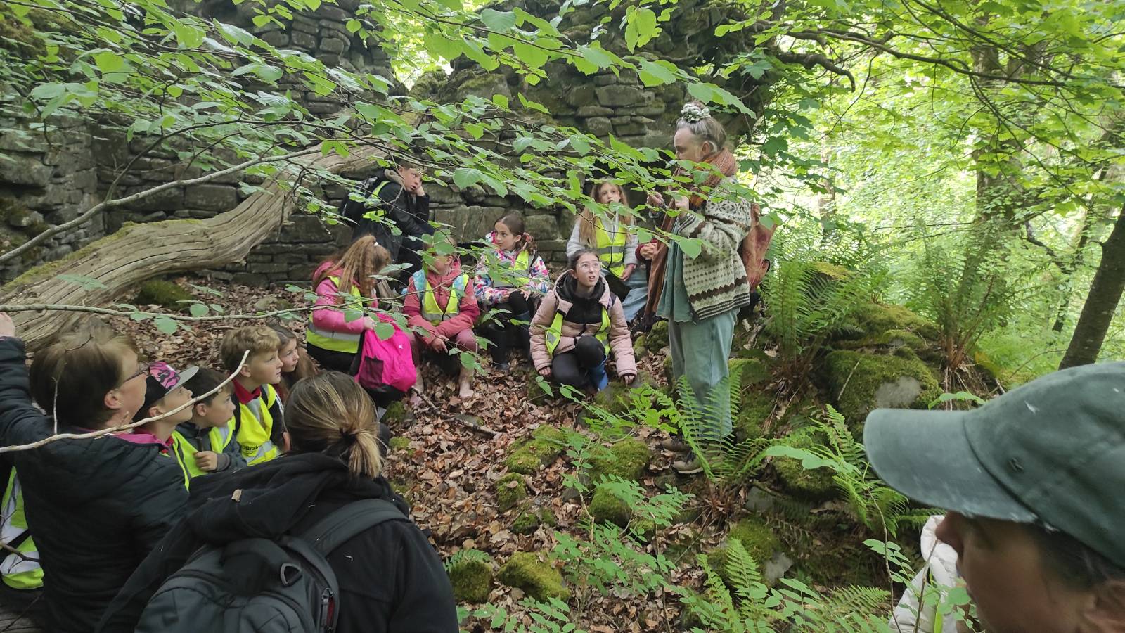 Storyteller animated, students with hi-vis jackets, seated in ruined cottage
