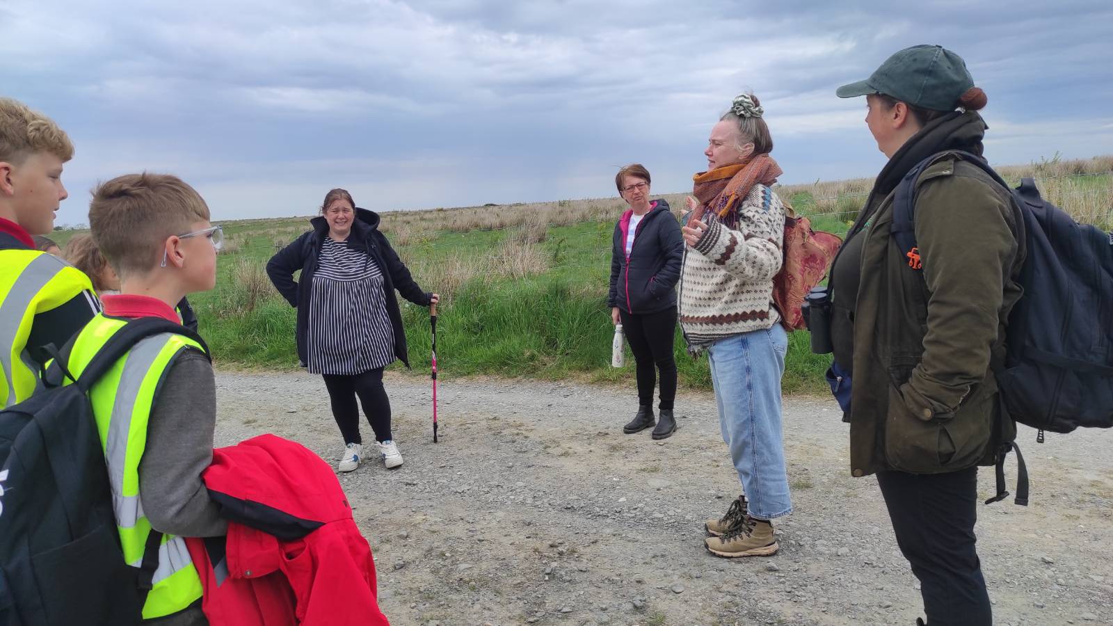 Four adults standing on dusty track next to field, talking to students