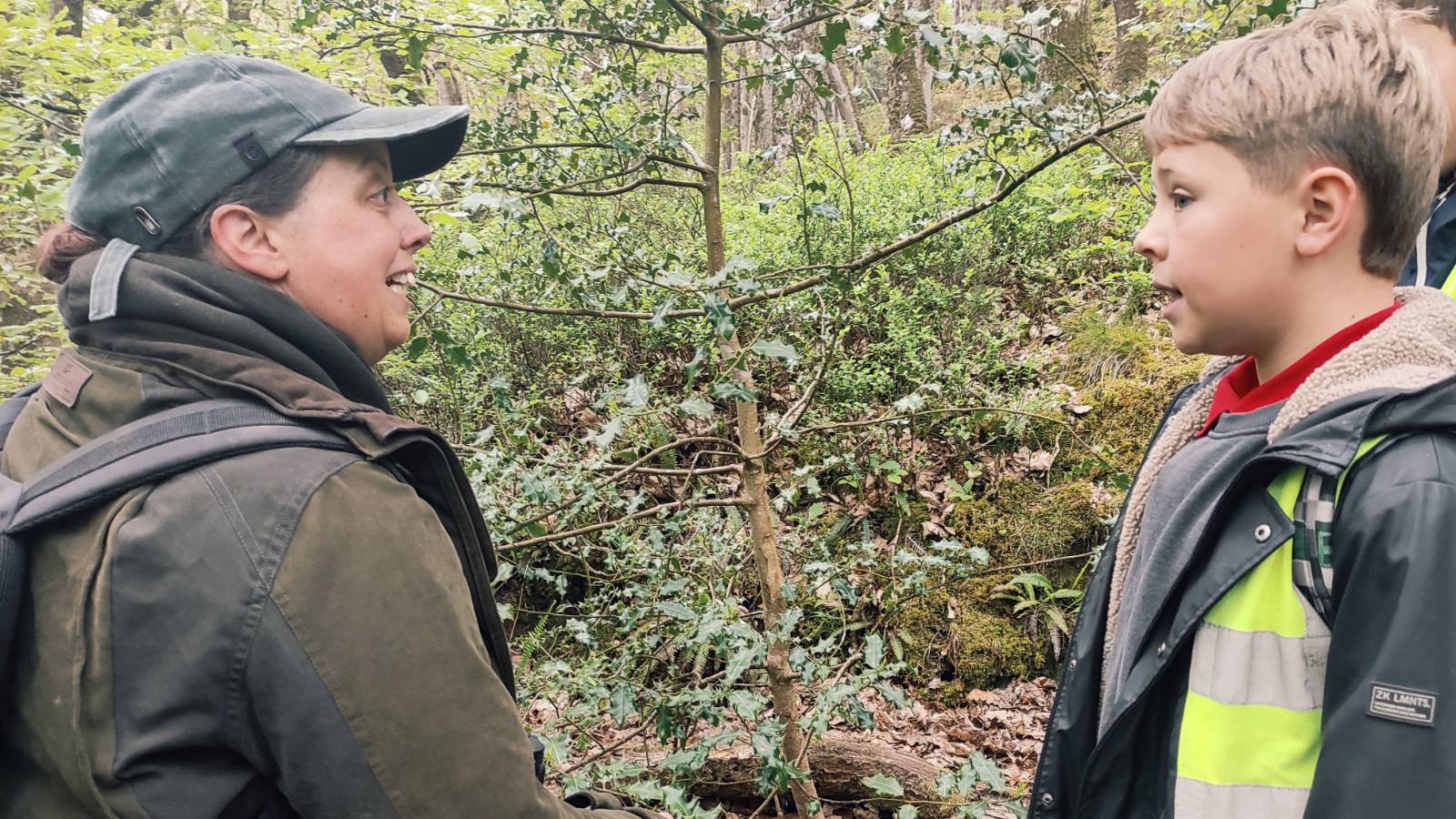 Ecologist with cap talks to student with hi-vis jacket about a prickly green shrub they're standing next to