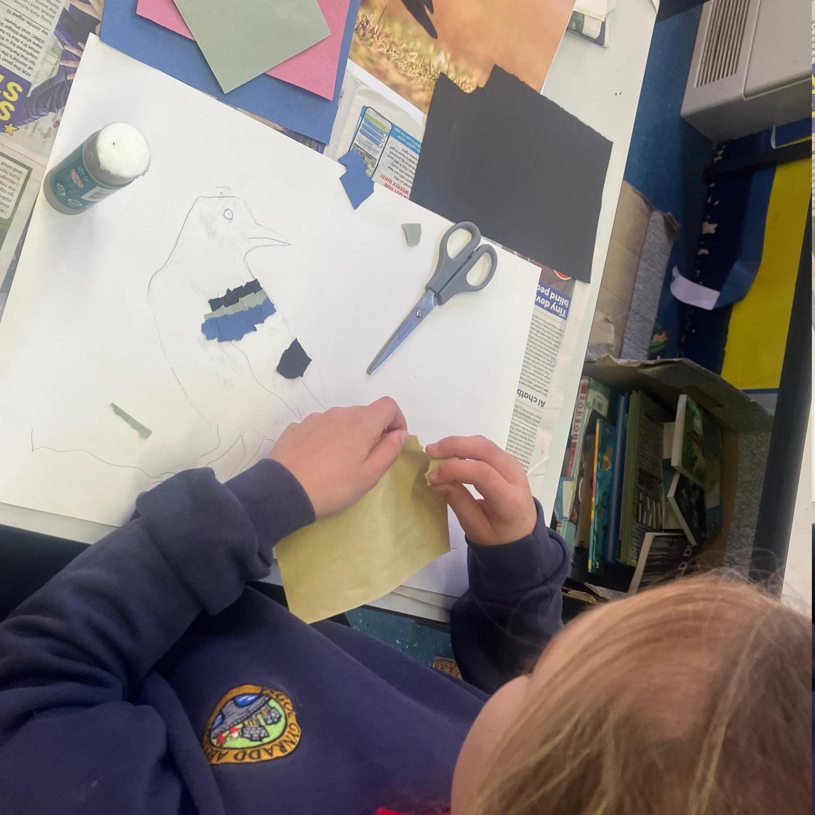 Student tearing yellow card, about to add to collage on top of pencil drawing of bird