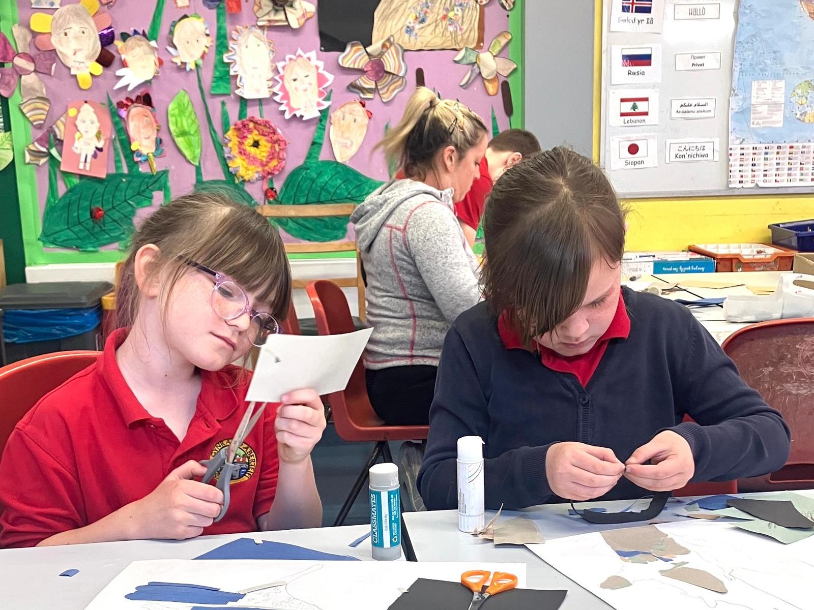 One student cutting card with scissors, another gluing card on a collage