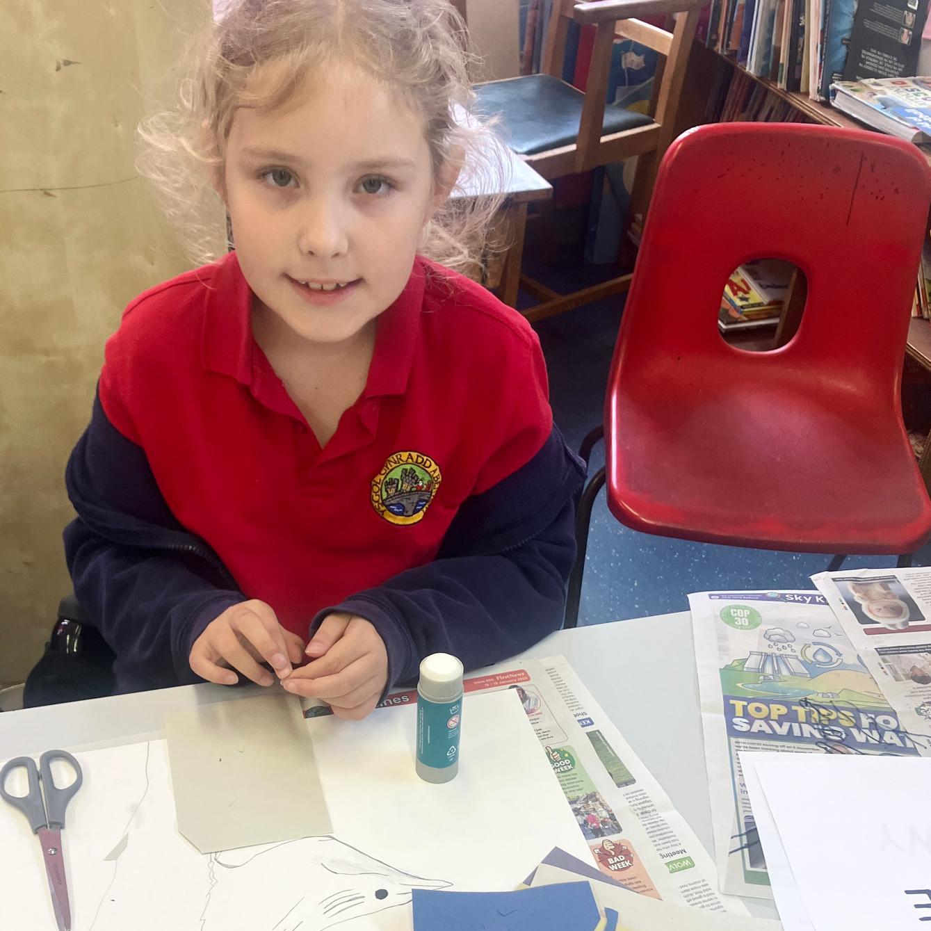 Student at desk looking at camera, with scissors, glue stick, and lots of paper
