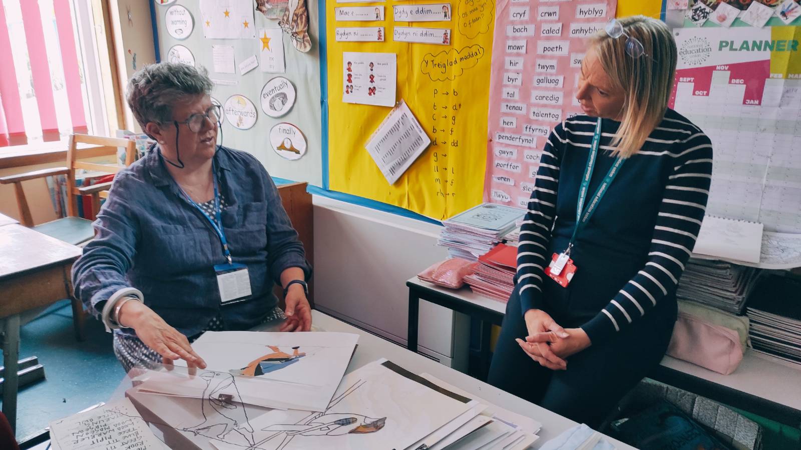 Artist sat at desk with collage of bird, talking with teacher leaning against table