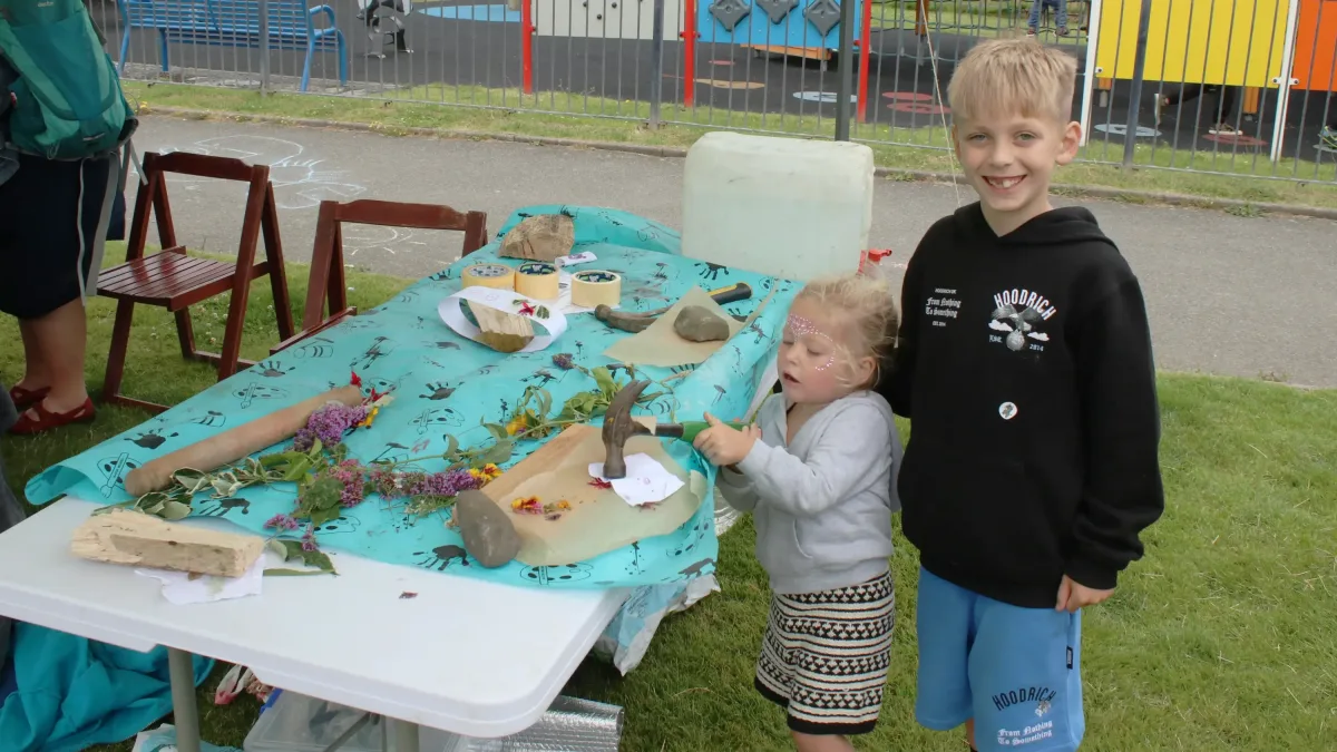 Two young kids, one under 5 with a hammer bashing something on an interesting table strewn with interesting things