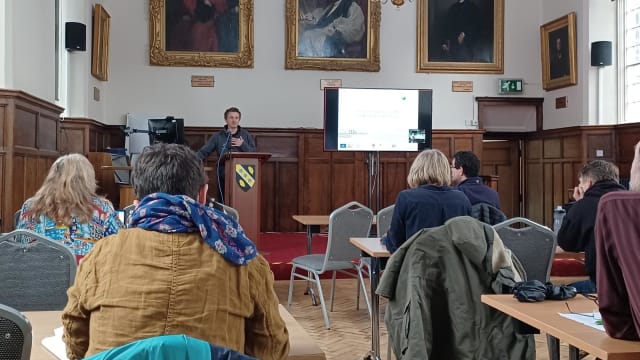 Meeting being held in university room with wood panelling and portraits on the wall, speaker at a podium with a crest of arms