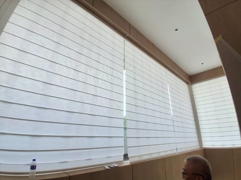White roman blind lowered over a sunlit window above a built-in wood bench with a water bottle and gloves nearby