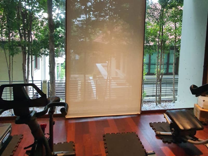 beige sunscreen roller blinds lowered between two glass walls in a home gym, trees visible through the mesh weave