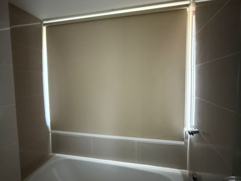beige blackout roller blind fully lowered over a bathroom window above a built-in bathtub, blocking any outside view