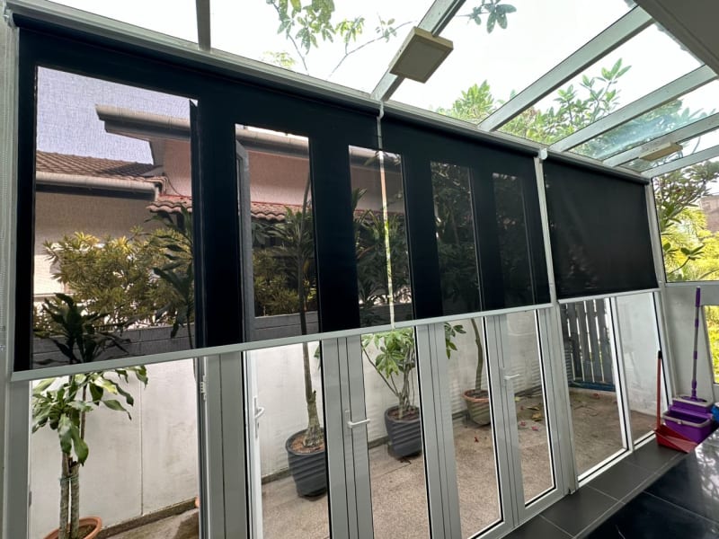 charcoal sunscreen roller blinds lowered across a glass courtyard roof and walls, rooftops visible through the mesh