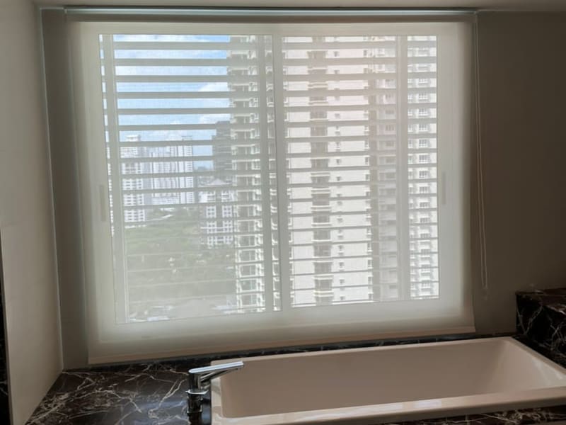 white sunscreen roller blind lowered over a window above a freestanding tub, high-rise towers visible through the mesh