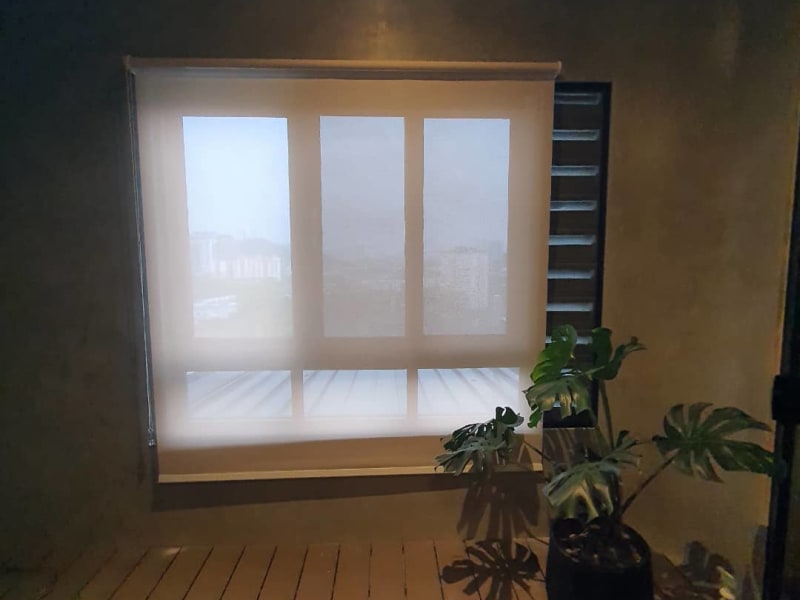 beige sunscreen roller blinds lowered on a balcony window at night, city rooftops and lights visible through the mesh
