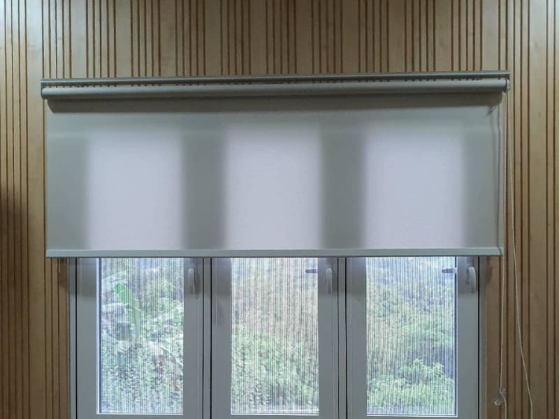 beige dim-out roller blind partly lowered over a triple window, greenery visible through the uncovered lower panes