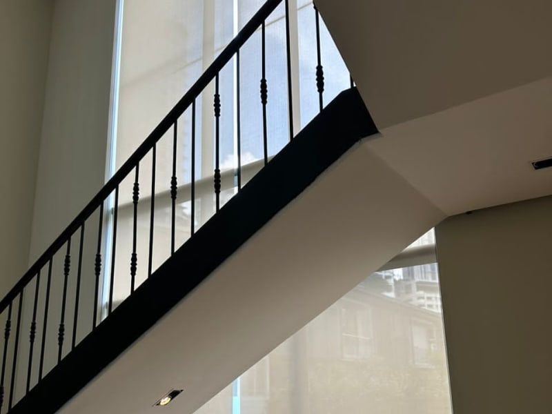 beige sunscreen roller blinds lowered over a tall stairwell window, a high-rise building faintly visible through the mesh