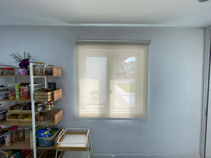 cream light-filtering roller blinds lowered over a kitchen pantry window with a neighboring house softly visible beyond