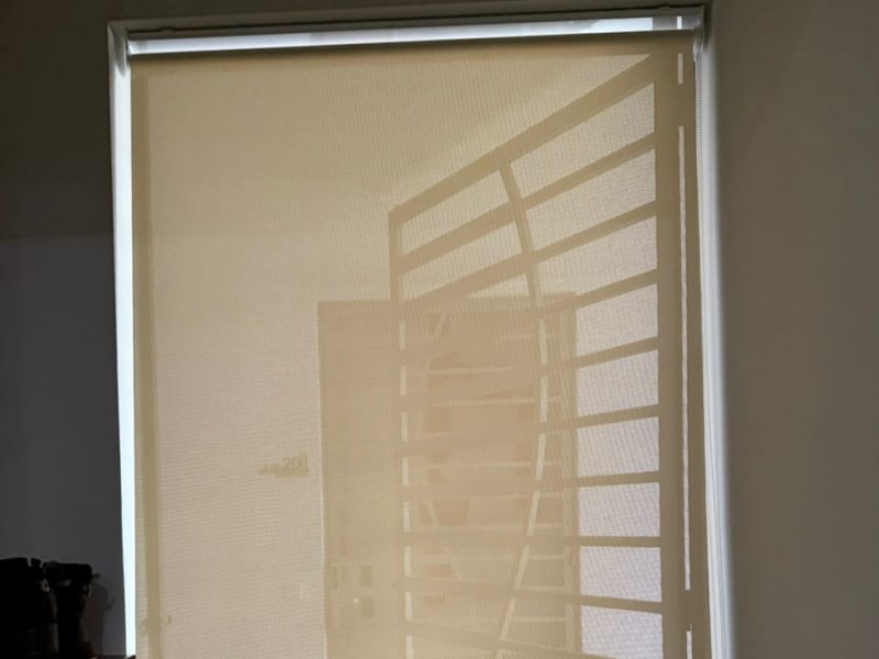 cream sunscreen roller blinds lowered over a foyer window, the outline of a spiral staircase visible through the mesh