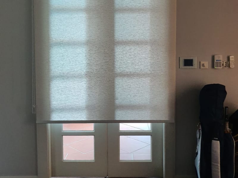 beige sunscreen roller blinds lowered over a glass double door, the door frame and floor tiles visible through the mesh