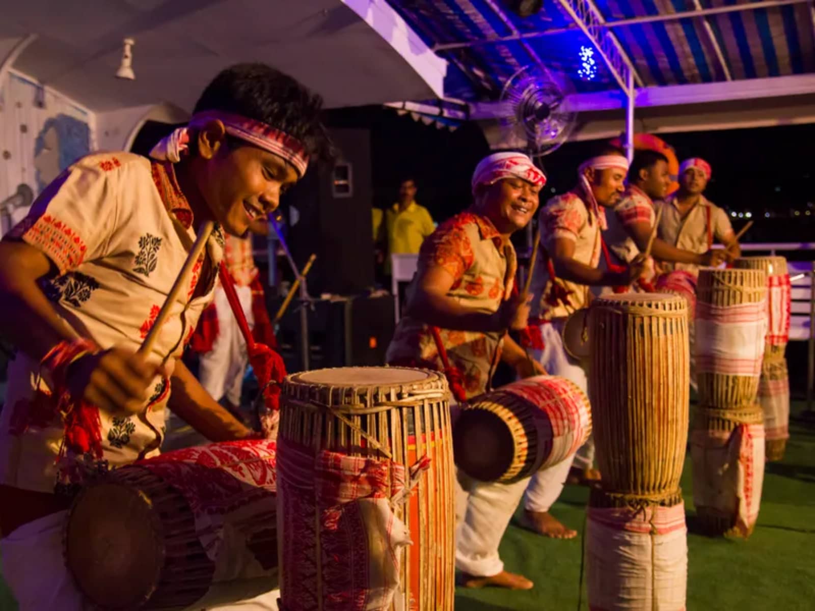 Assamese folk percussionists wearing traditional headbands playing dhol drums on the wooden boat deck.