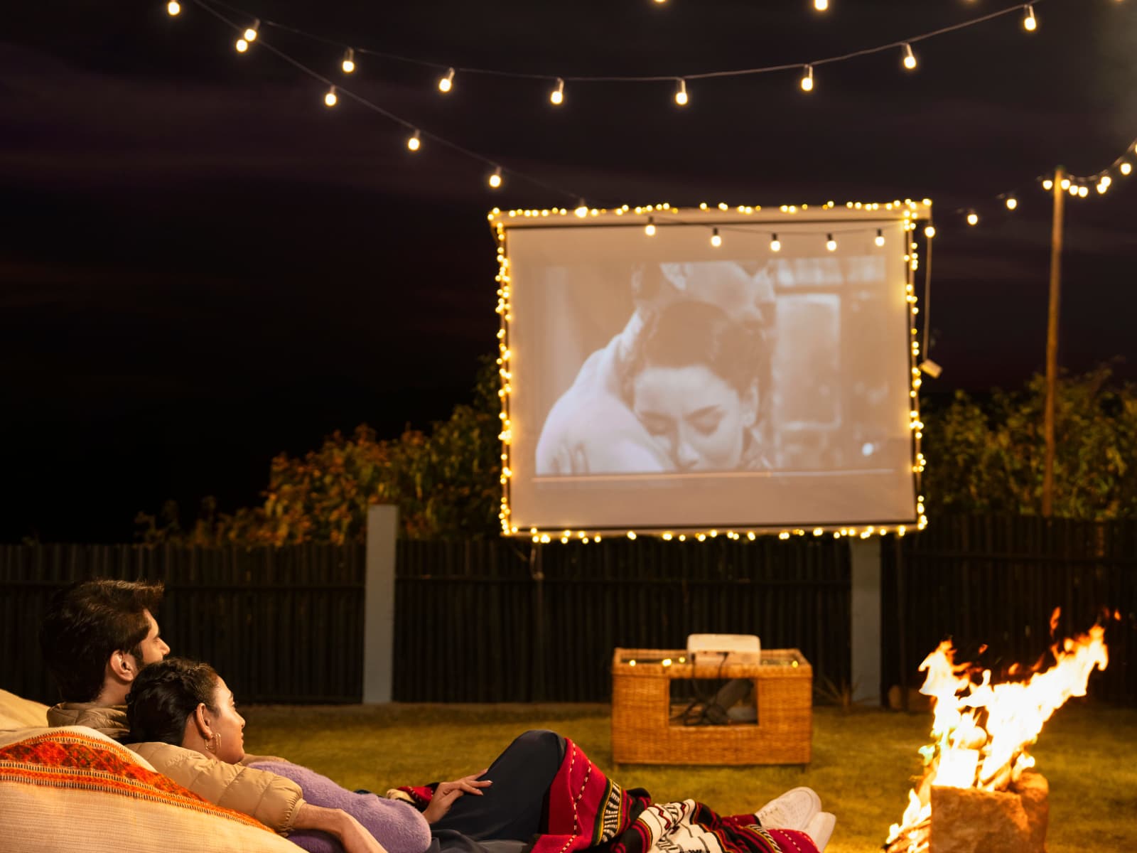 Guests on beanbags under string lights watching an open-air cinema screen next to a crackling bonfire pit at night