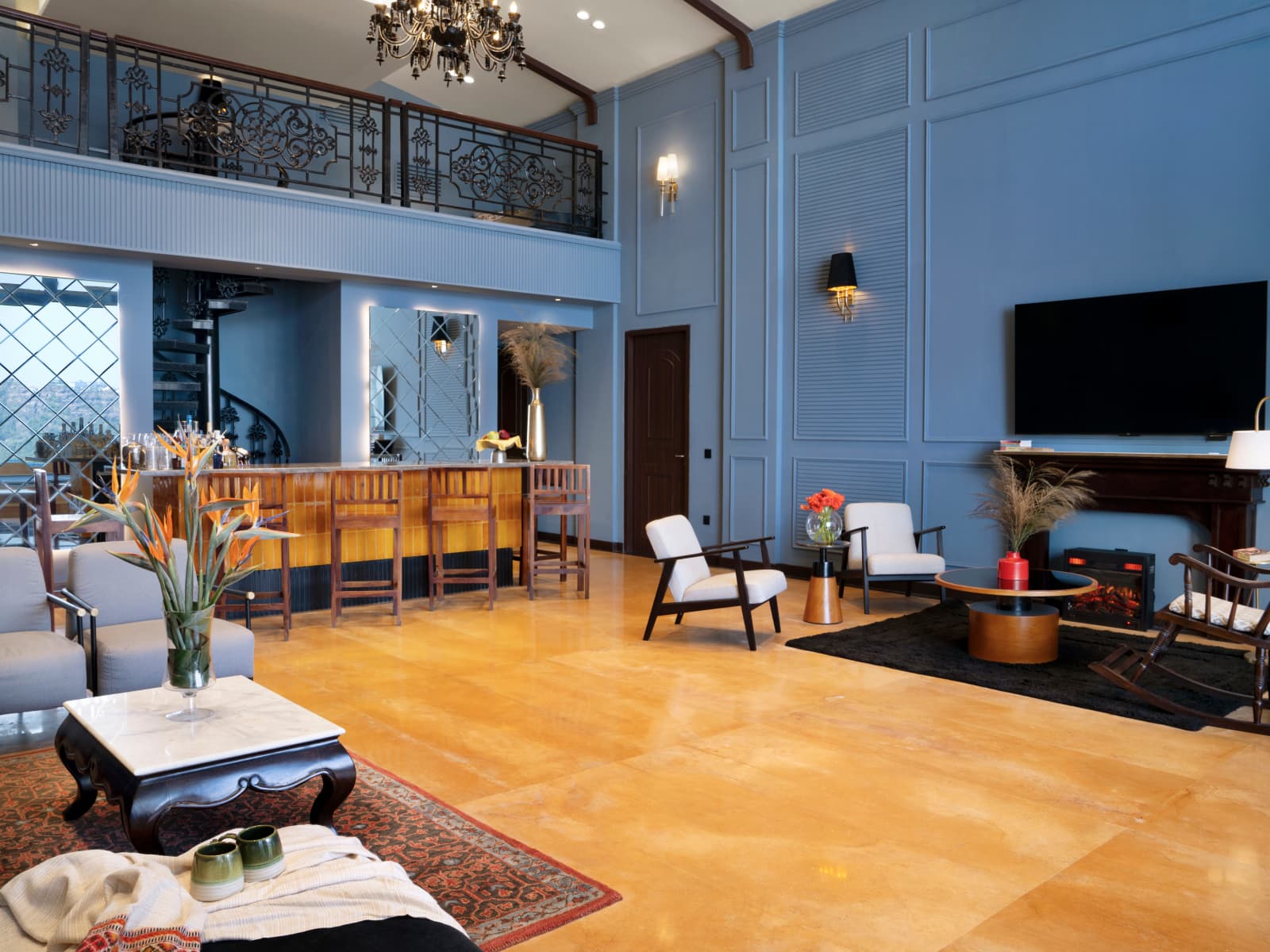 Double-height resort lounge and bar with powder blue walls, mezzanine balcony, burning fireplace, curved bar counter, and spiral stairs