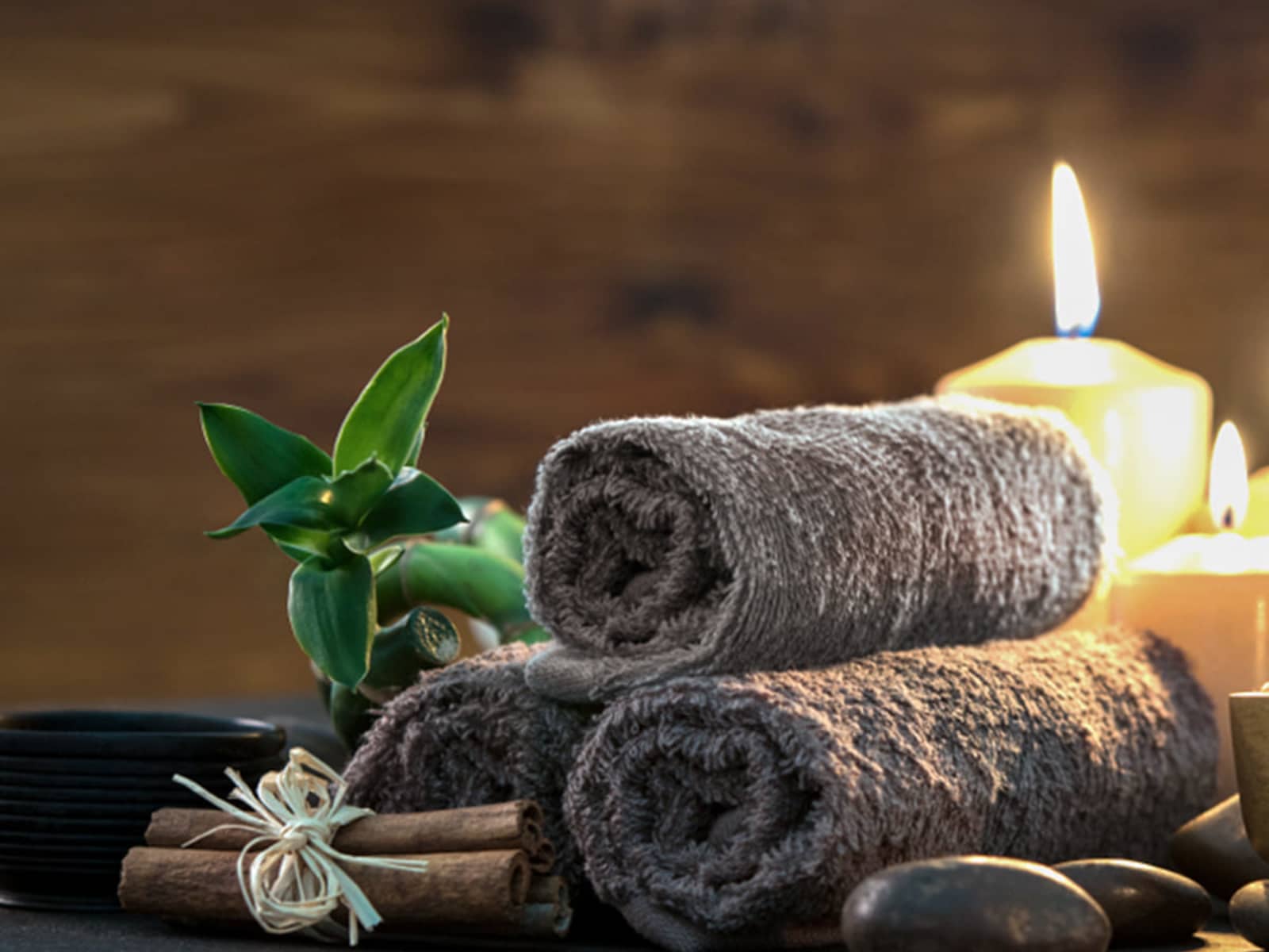 Close-up of spa wellness products including rolled brown towels, lucky bamboo plant, lit candles, cinnamon sticks, and massage stones