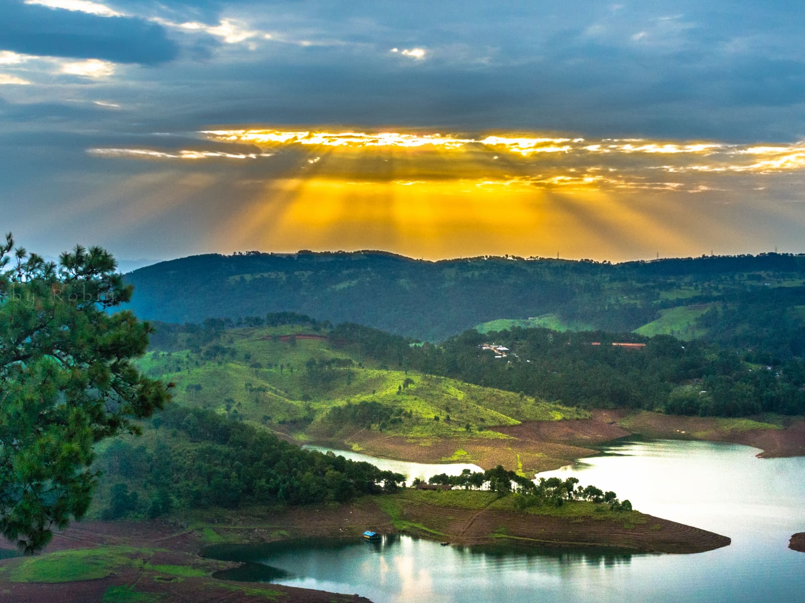 Crepuscular sunbeams radiating through clouds over Umiam Lake and surrounding Khasi hills at sunset.