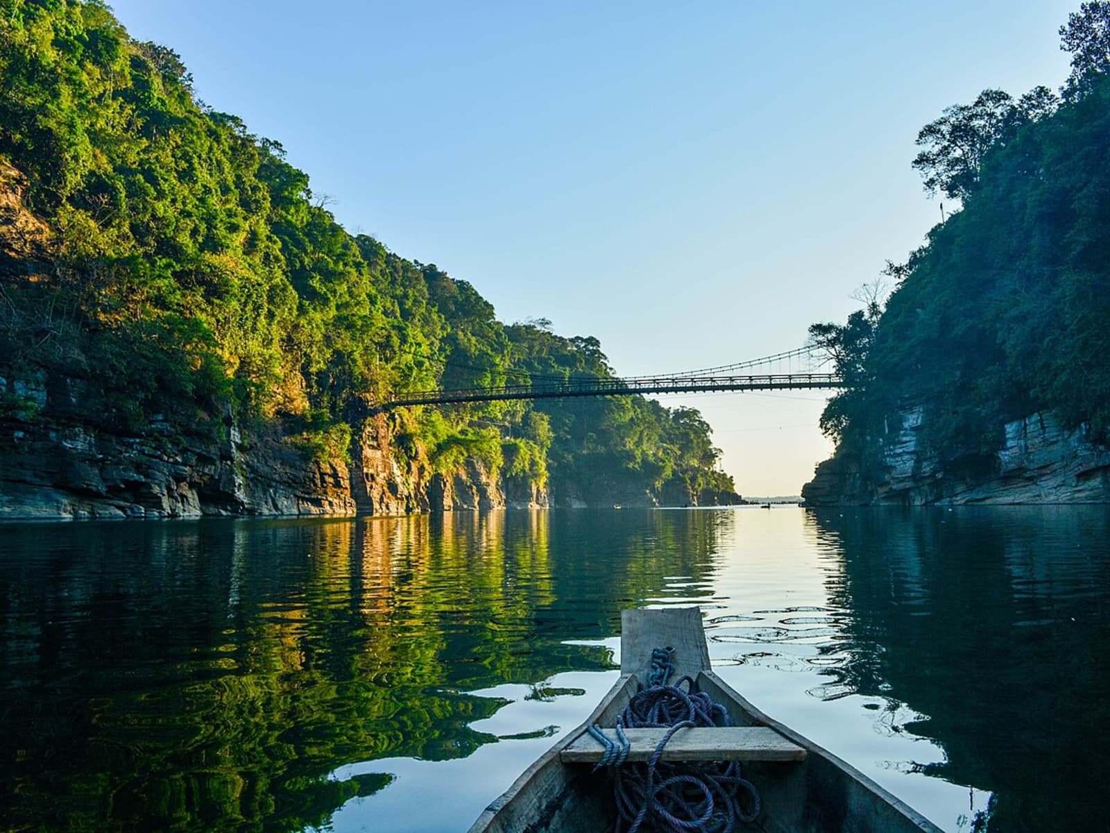 Wooden canoe prow pointing toward the historic Dawki suspension bridge across forested canyon cliffs.