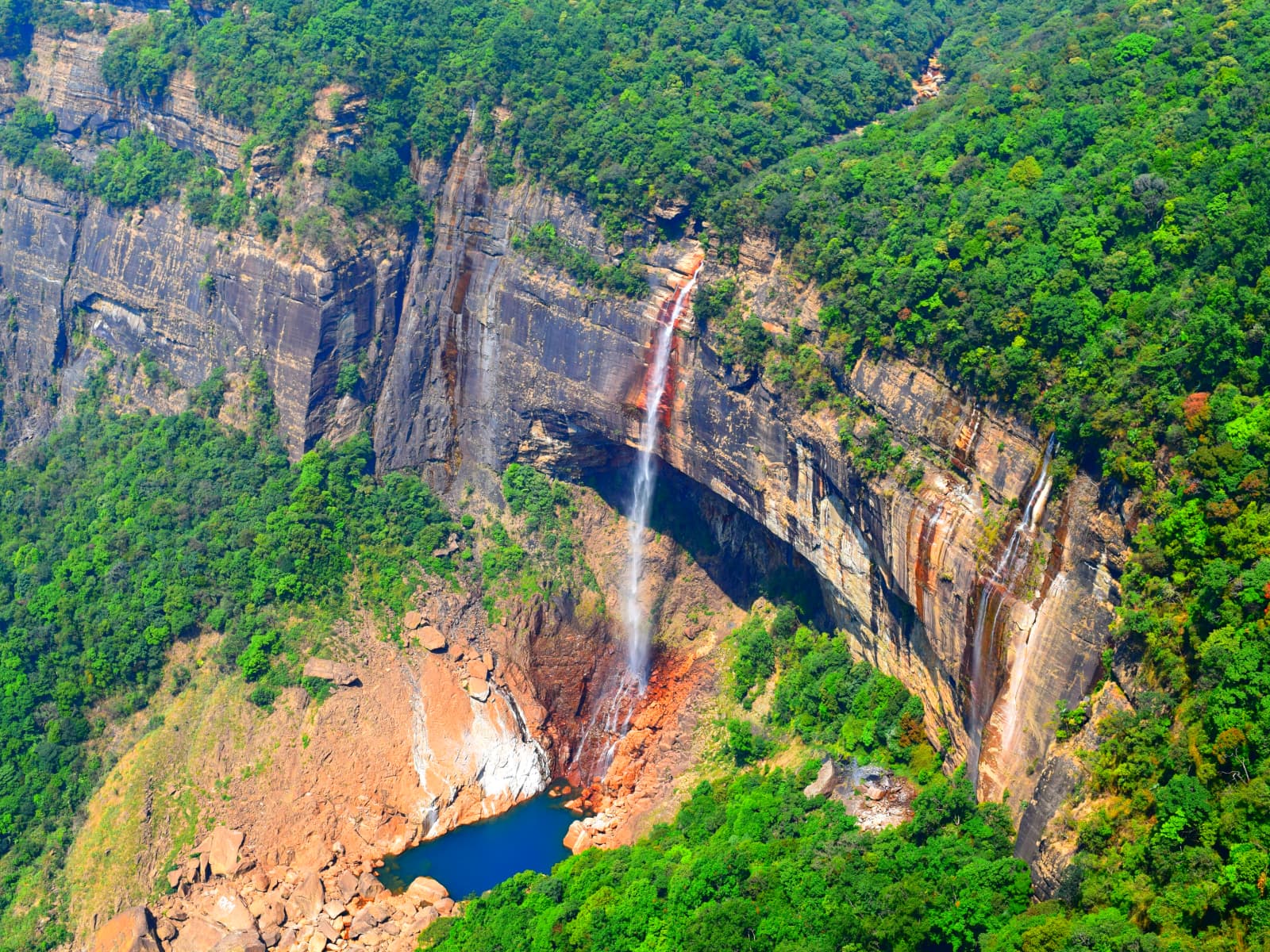Broad canyon gorge amphitheater enclosing Nohkalikai Falls and forested mountain slopes.