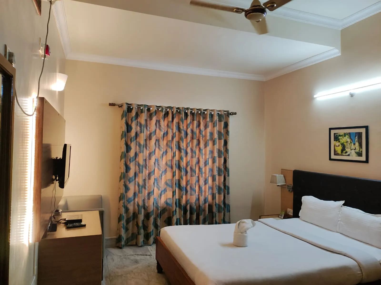 Deluxe King Room AC with king bed, folded towel swan art, window drapes, wall TV, and ceiling fan.