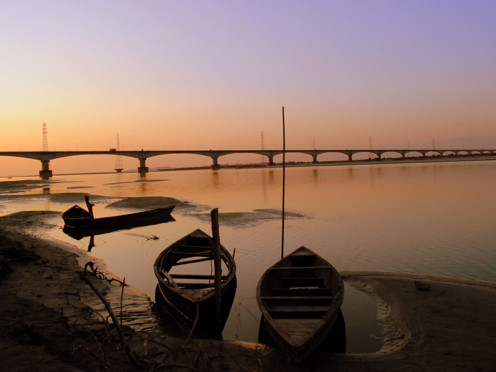 Wooden country boats moored along the Bhomoraguri riverbank silhouette against a fiery sunset and distant bridge.