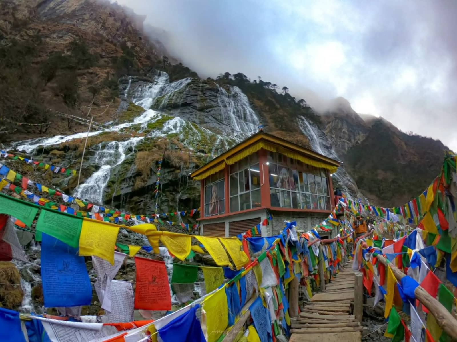 Wooden walkway lined with Buddhist prayer flags leading toward a glass prayer shrine before cascading waterfalls.