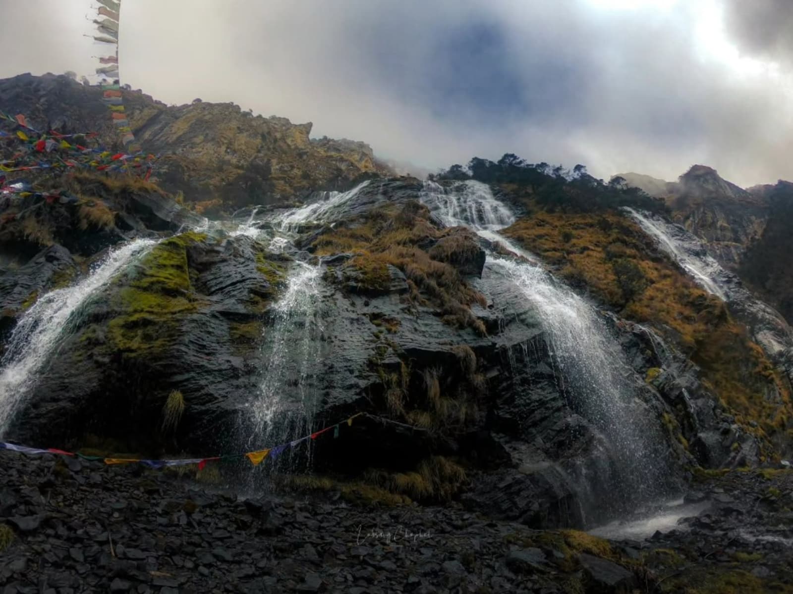 Water cascades down a dark mossy granite cliff into a stony stream bed hung with prayer flags at Chumi Gyatse.