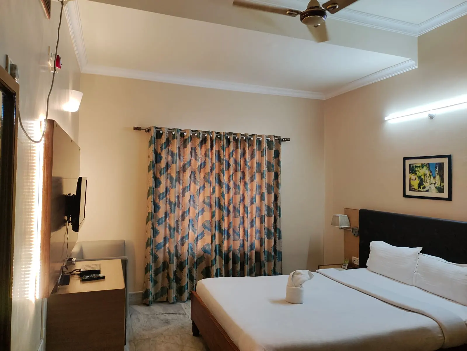 The Deluxe King Room AC features a padded king bed dressed in crisp white linen with towel art, wall-mounted TV console, and blackout curtains.