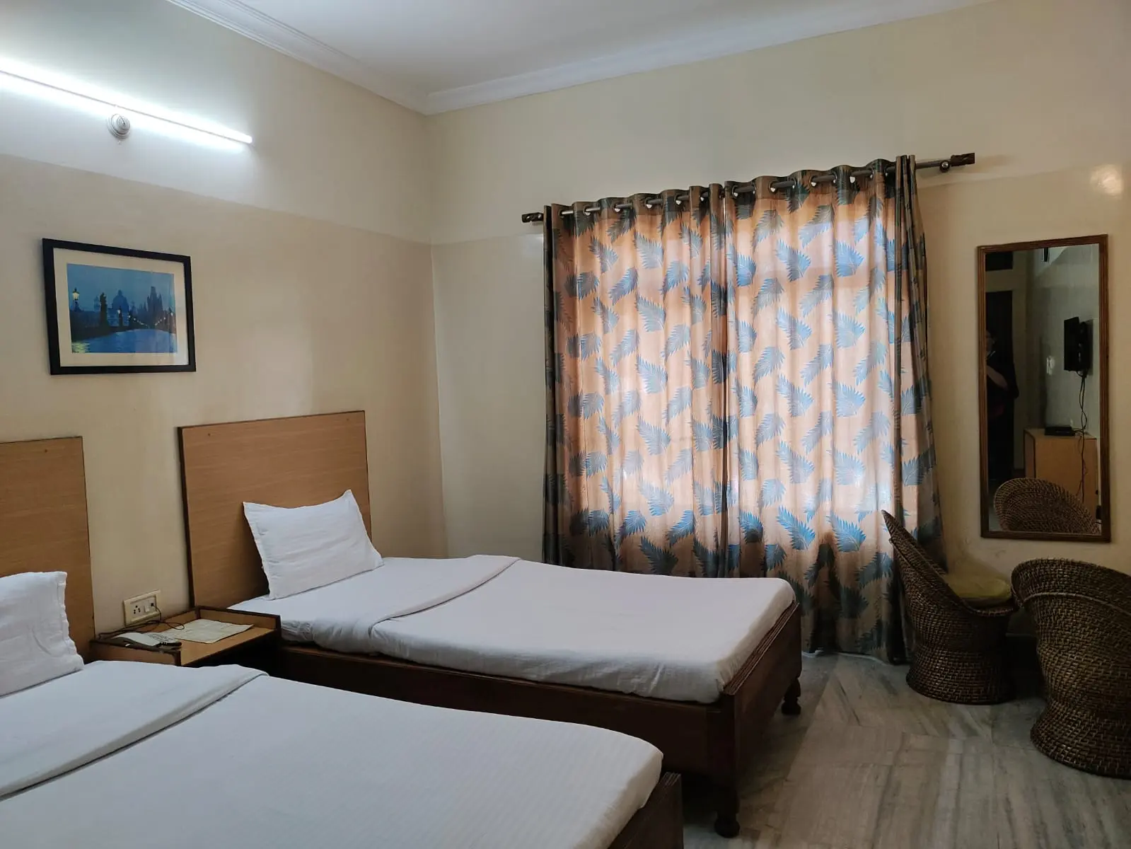 Budget-friendly Deluxe Non AC Room featuring two comfortable single beds, handcrafted cane wicker seating chairs, and large window curtains.