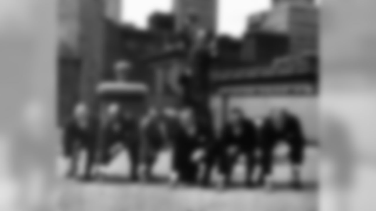 The coaching staff of the Titans of New York, American Football League, line up on rooftop for cameramen in New York, April 25, 1960. In background, Titan coach Sammy Baugh, former passing star of Washington Redskins, poses as if to pass. From left on the line are: Steve Sebo, assistant coach; John Steber, assistant coach; John Dell Isola, assistant coach; club president Harry Wismer; Dick Todd, assistant coach; and Hugh Taylor, assistant coach. The Titans, whose 1960 home schedule includes seven games, open at the Polo Grounds on Sept. 11. (AP Photo/John Lindsay)