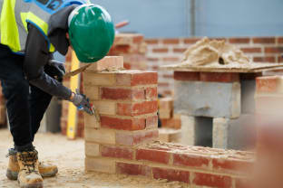 An apprentice in a green hard hat pointing a brick wall