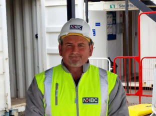 A man with white beard wearing a hi-vis jacket at a construction site, smiling at the camera