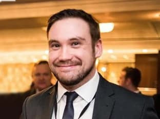 Josh Golding in black suit jacket and tie, with a white shirt - smiling at camera