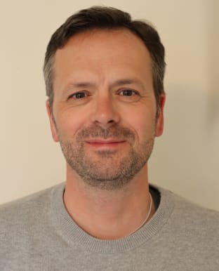 Adam Sadler smiling at the camera in a grey jumper