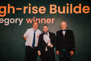 Lyam Gunner smiling at the camera with his Pride in the Job 2024 award alongside CEO Steve Woods and host Al Murray